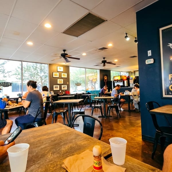 Diners enjoy meals at tables in a casual restaurant with large windows and a vintage poster.