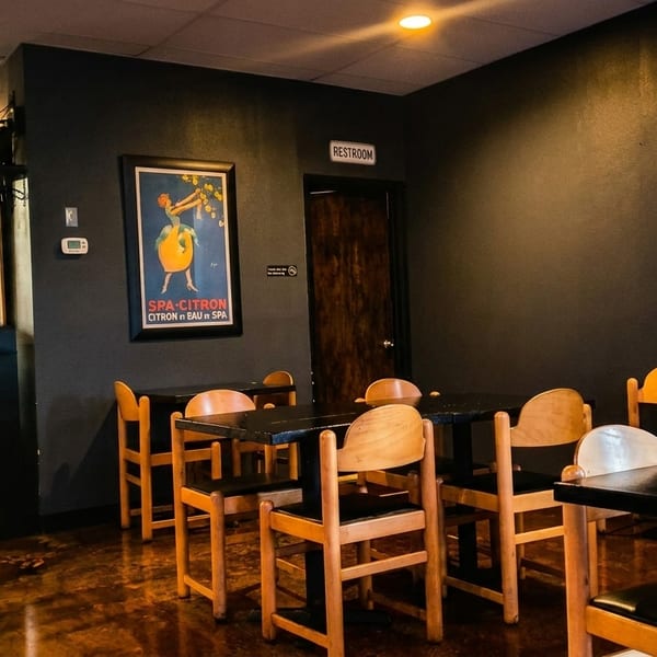 Restaurant interior with dark walls, wooden tables and chairs, and vintage posters, with a kitchen in the background.