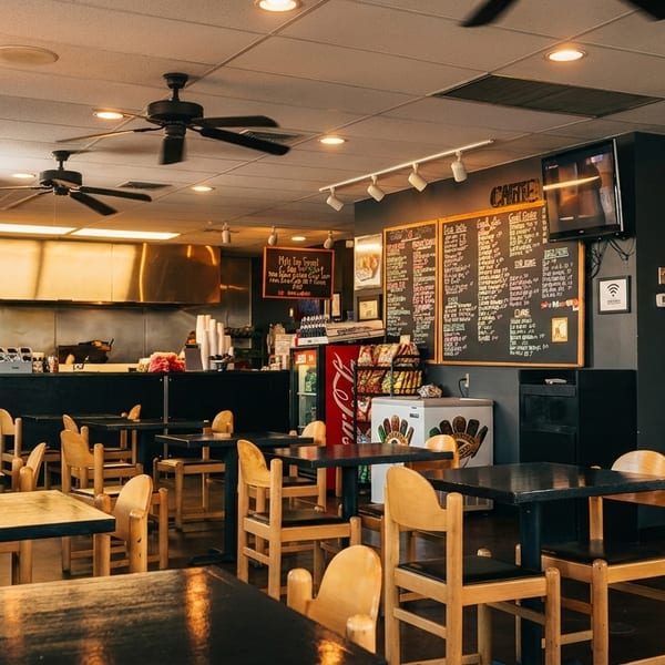 Interior of a casual restaurant with dark tables, wooden chairs, and a chalkboard menu.