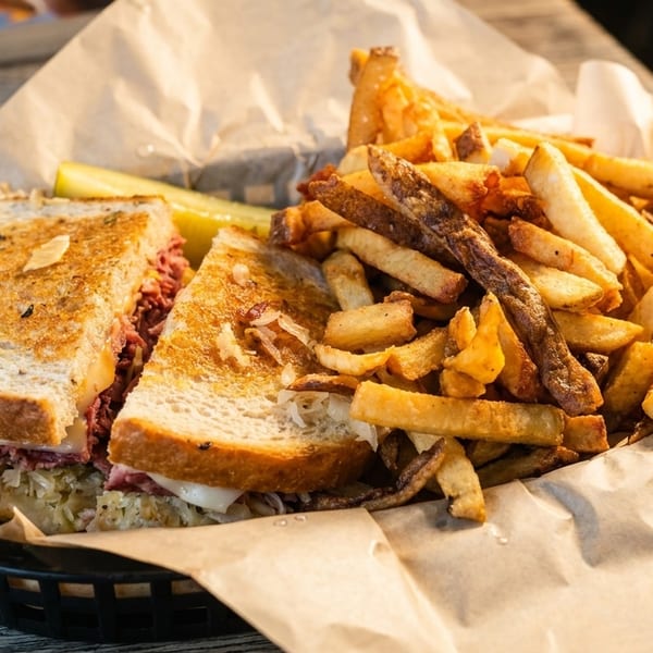 A reuben sandwich with fries and a pickle spear served in a basket on a wooden table.
