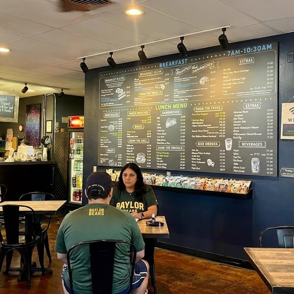 Customers seated at tables in a casual restaurant with a large menu board on a dark blue wall.